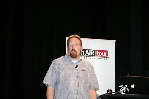 A speaker standing on stage in front of a banner for the AIR Tour Europe 2008. He has a short beard and wears a plaid shirt, gesturing while addressing the audience. A laptop is visible on a table beside him.