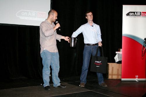 Two presenters are on stage at an event, one holding a microphone, and the other holding a container and a tote bag. A branded banner and projector screen are visible in the background.