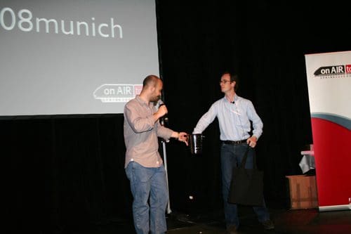 Two men standing on a stage at a conference. One man, in a light shirt, is holding a microphone while the other, wearing a blue shirt, receives a small bucket. A banner with 'on AIR' is visible in the background.
