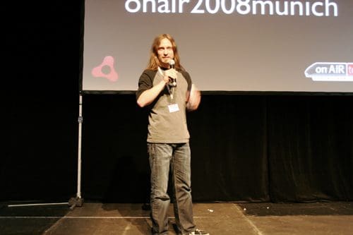 A speaker with long hair stands on a stage, holding a microphone and addressing an audience at a conference. A large screen behind him displays the text 'onair2008munich'. The stage features dark curtains and a simple setup.