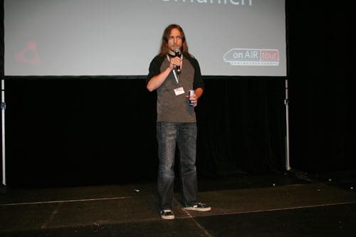 A speaker stands on a dimly lit stage, holding a microphone in one hand and a beverage in the other. He wears a black shirt with gray sleeves and jeans, speaking to an audience in front of a screen displaying the event logo.
