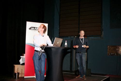 Two presenters at a conference standing behind a high table. One speaker is looking at a laptop, while the other gestures and speaks to the audience. A banner in the background displays the text 'on AIR'. The setting is a dimly lit stage.