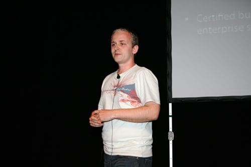A speaker stands on stage in a casual white t-shirt with graphic designs, addressing an audience. Behind him, a projection screen displays information about business and enterprise.