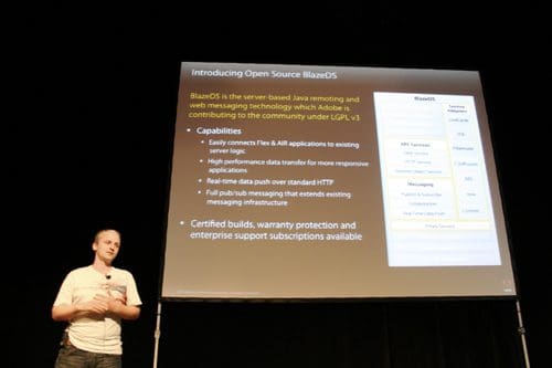 A presenter speaking at a conference, with a visual projection behind him displaying information about Open Source BlazeDS, including its capabilities and features, in a dimly lit auditorium.