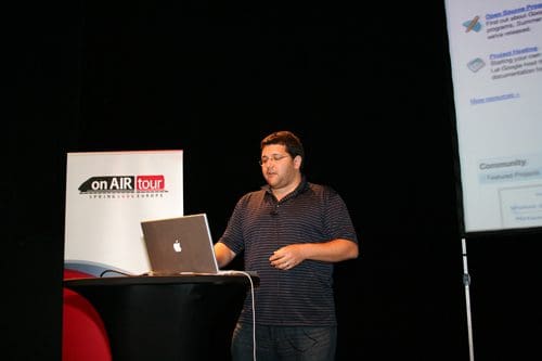 A man presenting at a speaking event, standing behind a podium with a laptop. He is wearing a striped shirt and appears engaged with the audience. A banner in the background reads "on AIR tour".