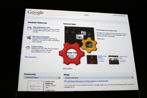 A computer screen displaying the Google homepage featuring developer resources, community projects, and featured news. Two colorful gears are superimposed in the center, symbolizing development and collaboration.