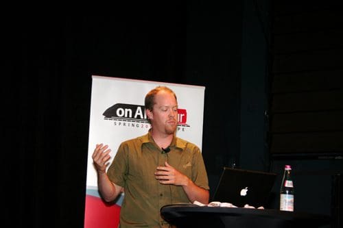 A speaker gesturing with one hand while discussing at a presentation, standing in front of a branded backdrop. A laptop is placed on a table beside a bottle of water.