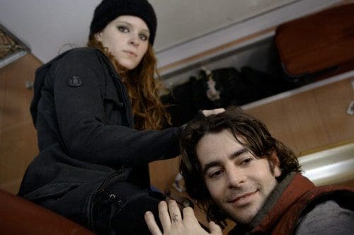 A woman with long, wavy red hair, wearing a black hat and dark clothing, looks intently at the camera while sitting on a low surface. A man with dark, tousled hair sits below her, smiling and gazing at her.
