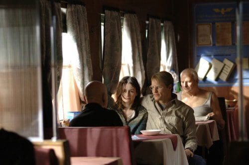 A cozy diner interior with warm lighting, featuring a man and a woman engaged in conversation across a table, while another woman sits alone in the background. The decor includes curtains and framed certificates on the wall.