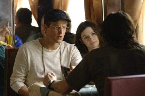 A man wearing glasses and a cap engages in conversation across a table with a woman, who listens attentively. They are seated in a cozy restaurant with warm lighting and soft furnishings.