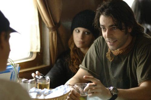 A man with tousled hair and a green shirt, engaged in conversation, gestures with his hands. A woman in a black beanie and long hair sits nearby, listening intently. They are at a table with drinks and an inviting atmosphere in a cozy setting.