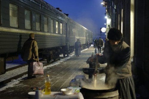 A woman stirring a pot of food on a snowy train station platform, with train cars in the background. Passengers walk by, some carrying bags, in the dim light of early evening.