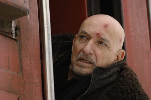 A middle-aged man with a bald head and a rugged appearance peeks out of a train window. He has a worn expression and noticeable marks on his forehead, suggesting he has been through a struggle.