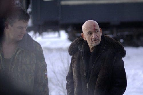 Two men stand outdoors in a snowy landscape. One man, wearing a dark fur coat, looks solemn with a visible bruise on his forehead. The other man, dressed in a military-style jacket, appears concerned as they engage in conversation near a train.