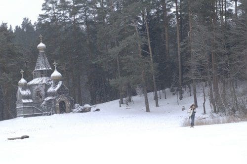 A serene winter landscape featuring a small ornate church with onion domes, surrounded by snow-covered trees. A person stands in the distance, dressed in winter attire, against a backdrop of a tranquil snowy field.