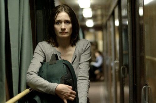 A woman with shoulder-length brown hair stands in a dimly lit train corridor, holding a green backpack. She has a concerned expression, looking directly at the camera as the reflection of other passengers is seen in the background.