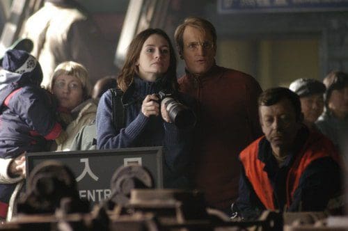 A woman holding a camera stands beside a man in a busy train station. They look intently at something in the foreground, while a crowd, including a person with a child, gathers around them. The background features signs in a different language.