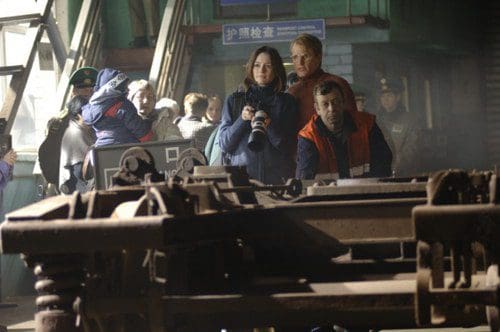 A woman with a camera stands in front of a train track, examining the scene, while a man beside her observes intently. In the background, a small crowd watches, with officials in uniforms present, set in a station with green walls.