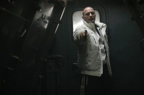 A man wearing a white puffy jacket stands in a dimly lit doorway, pointing outward. The scene is enclosed by dark metal, giving a tense atmosphere.