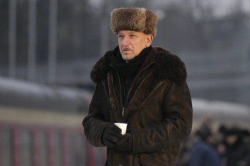 A man dressed warmly in a dark leather coat with fur trim and a furry hat stands at a train station, holding a cup in his gloved hand. His serious expression contrasts with the bustling background of the platform.