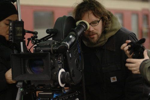 A crew member looking through the viewfinder of a professional film camera while another person adjusts the camera setup. The scene appears to be on a film set with a soft focus on the background.