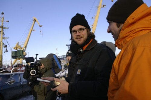 A man with glasses and a black beanie holds a clipboard, smiling and engaging with a colleague wearing an orange jacket. They are in a port area with cranes and a vintage car visible in the background.