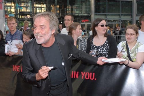A man in a black suit enthusiastically interacting with fans at a public event, holding a marker and smiling. A group of various fans, some holding items to be signed, stands behind a promotional banner.