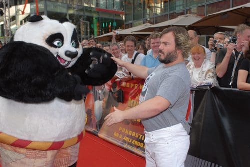 A giant panda character engages playfully with a man in a gray t-shirt and white pants on a red carpet, surrounded by an audience taking photos. The event atmosphere is celebratory, with people smiling and enjoying the moment.