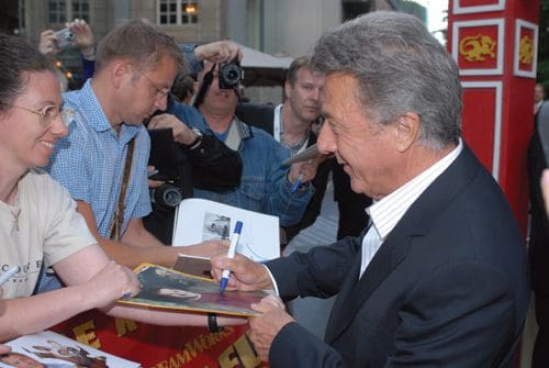 A well-known actor signing autographs for fans outside an event. A woman eagerly holds up a poster while he writes on it, surrounded by cameras and other attendees.