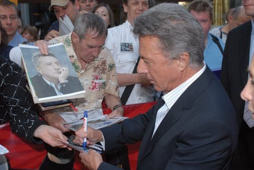 A well-known actor signing autographs for fans at an outdoor event. He is focused on a photograph being presented to him, surrounded by a crowd of enthusiastic supporters.