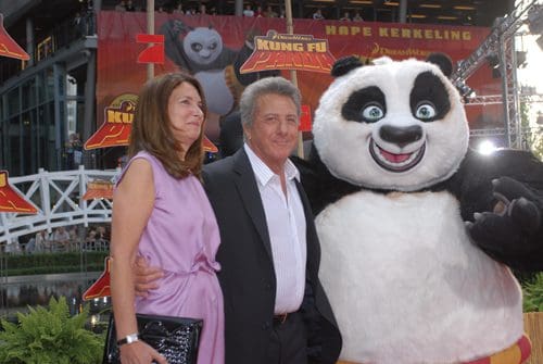 A couple poses together on a red carpet, smiling. They are joined by a large, friendly panda character, promoting the movie 'Kung Fu Panda,' with colorful banners in the background.