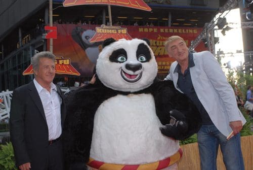 Two men stand beside a character dressed as a panda, at an outdoor event promoting a movie. The panda has a cheerful expression, and the background features colorful decorations related to Kung Fu.