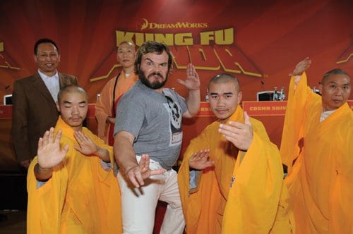A group of martial artists in orange robes are posing energetically at a promotional event for "Kung Fu Panda." In the center, a man with a beard and casual attire strikes a playful pose, flanked by smiling monks.