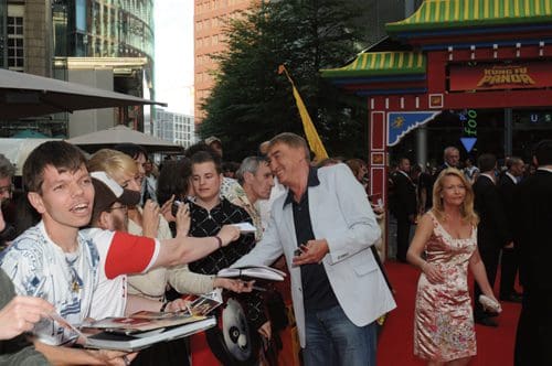 A lively red carpet event with fans gathering around celebrities. A man is reaching out for autographs while others chat. In the background, a colorful building adds to the festive atmosphere.