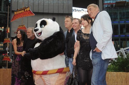 A group of five people posing with a large panda character at a movie premiere event, smiling and standing together on a red carpet in an urban setting.