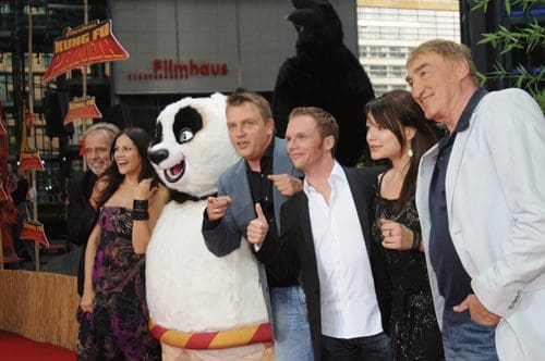A group of five people and a large panda mascot posing together on a red carpet at a promotional event. They are smiling, with the panda standing in the center. The background features colorful decorations related to the movie 'Kung Fu Panda'.