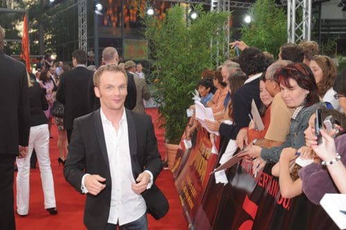 A man in a black suit jacket and white shirt walks along a red carpet at an event, smiling as he interacts with fans and photographers. The background features a lively crowd and greenery.