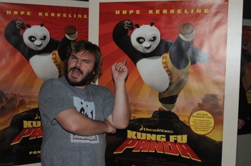 A man with long hair and a beard poses in front of a colorful 'Kung Fu Panda' movie poster featuring a cartoon panda performing a kick. The man is wearing a gray T-shirt with a panda design and is making a playful expression.