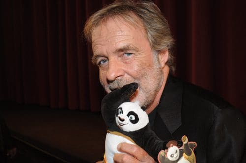 A smiling man with gray hair wearing a black suit holds a plush panda toy close to his face. The backdrop features a dark curtain, suggesting an event setting.