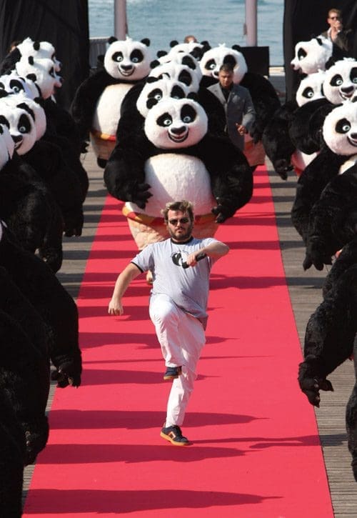 A man in a gray shirt runs energetically on a red carpet, flanked by numerous performers in panda costumes. The scene takes place outdoors near a body of water, creating a lively and whimsical atmosphere.