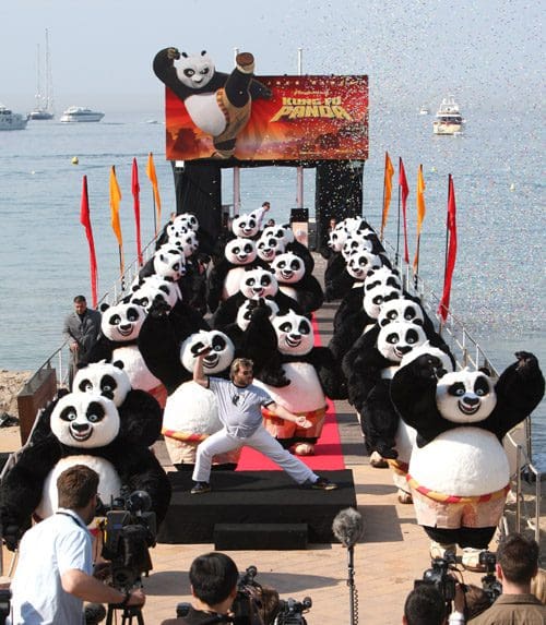A festive outdoor event featuring numerous people in panda costumes on a red carpet by the beach. A performer in white stands in front, striking a pose, while cameras capture the lively scene, surrounded by colorful flags and decorations.