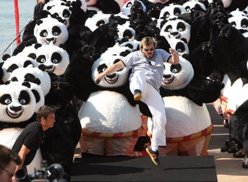 A man in a white outfit jumps forward amidst a crowd of large, plush panda costumes on a sunny day. Two men, one in black and another in a white outfit, stand on a stage, surrounded by cheerful panda figures.