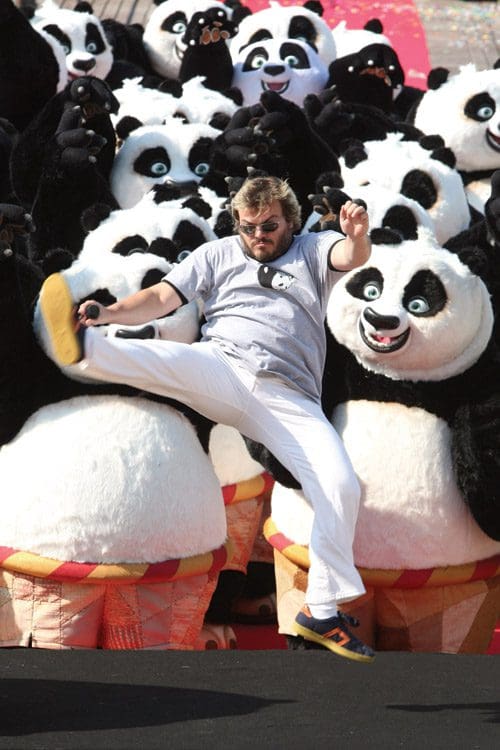 A man wearing a light gray shirt and white pants is slipping in front of a large group of cartoonish panda plush toys, each with distinct facial expressions, set against a vibrant background.