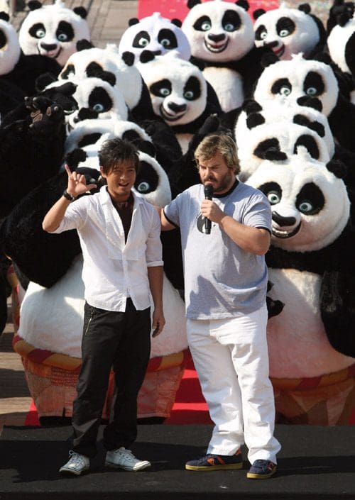 Two men stand on a stage speaking into a microphone, with a crowd of costumed panda characters behind them. One man wears a white shirt, and the other has a gray t-shirt and white pants, set in a festive outdoor setting.