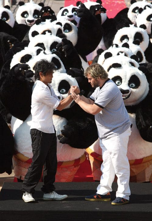 Two men, one in a white shirt and black pants and the other in a gray shirt and white pants, engage in a playful arm-wrestling match on a stage. Behind them, numerous panda mascots create a festive atmosphere.