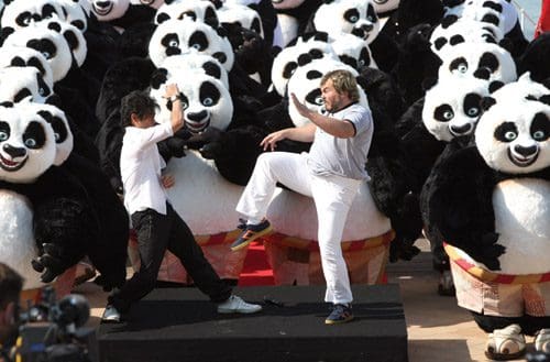 Two performers, one in a black suit and the other in a gray shirt, are playfully mimicking each other on a stage surrounded by numerous people dressed in panda costumes. The backdrop features a lively crowd of pandas.