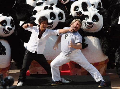 Two men engage in a comical action pose in front of a crowd of large panda mascots during a promotional event. The men are laughing, one wearing a black shirt and the other in a gray shirt, with a playful, animated atmosphere surrounding them.
