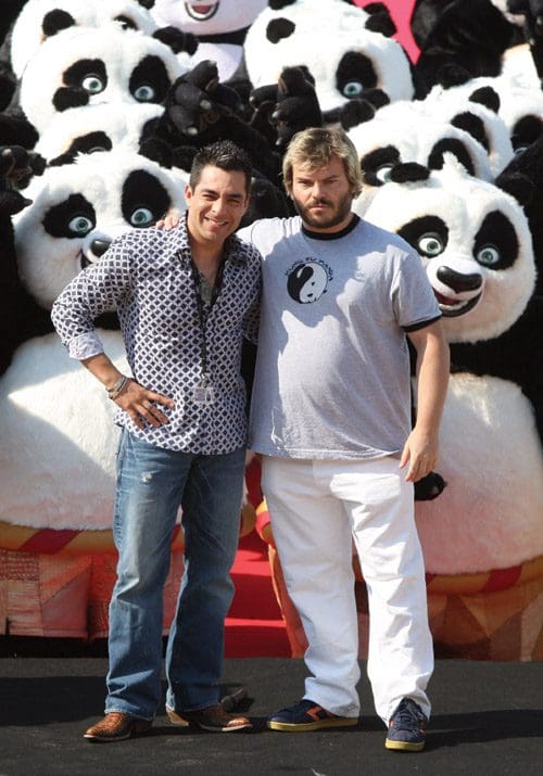Two men posing together in front of a large group of panda mascot figures. One man is wearing a patterned shirt and jeans, while the other is dressed in a T-shirt and white pants. The setting appears festive with colorful decorations.