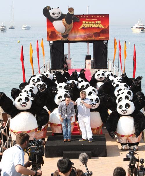 A large group of people in panda costumes waving their arms, standing on a stage set up by a waterfront. Two men, one in casual attire and the other in a white outfit, are addressing a crowd of photographers and onlookers. A large banner promoting "Kung Fu Panda" is in the background.