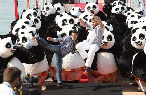 Two men perform martial arts poses on a colorful stage surrounded by numerous large panda mascots. The setting appears festive, with bright decorations and water in the background.